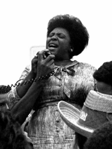 Fannie Lou Hamer speaking into a handheld microphone and surrounded by people