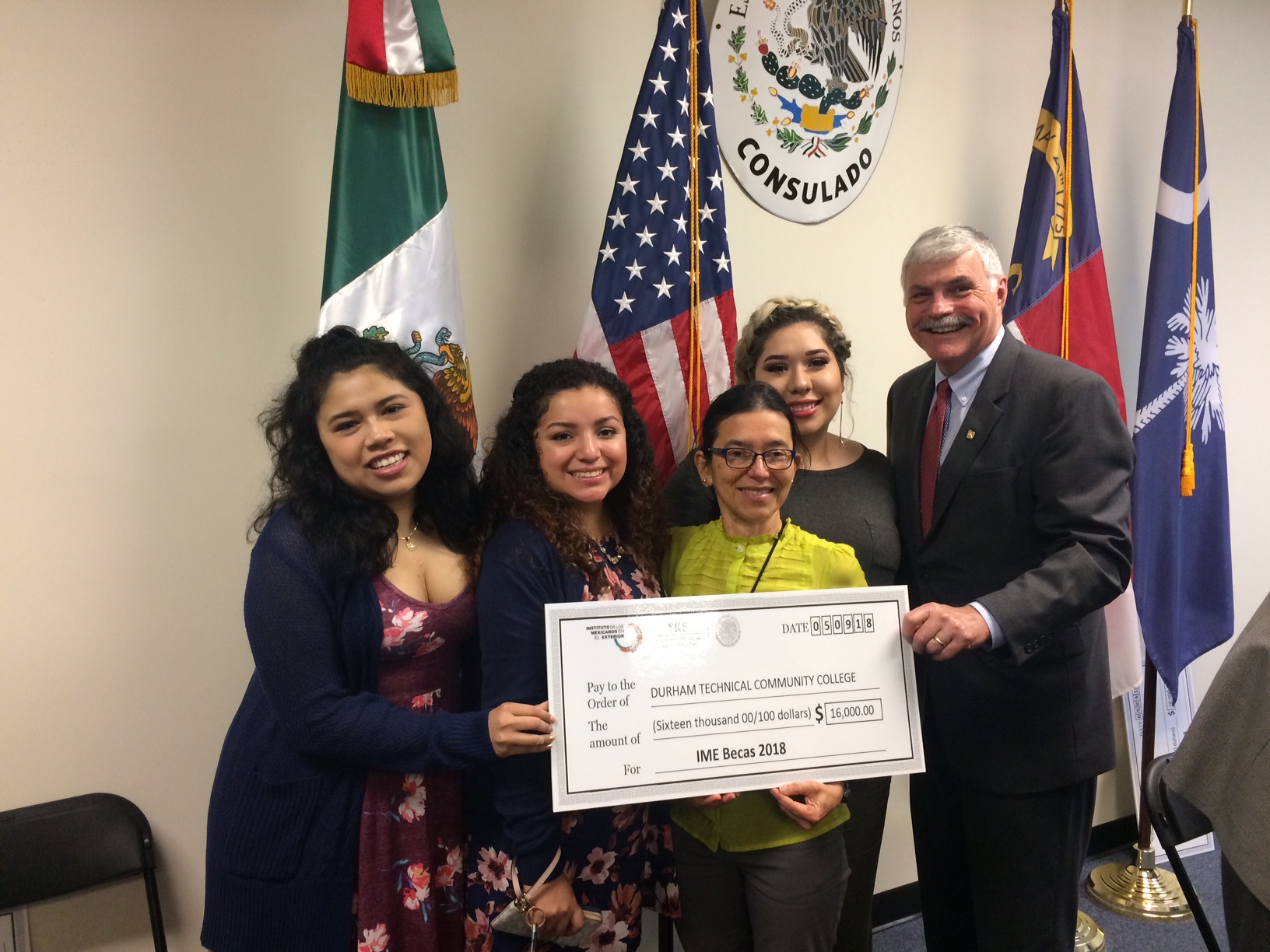 Four students and Dr. Ingram holding check in front of four flags, including Mexico's flag, American flag, North Carolina Flag and South Carolina Flag