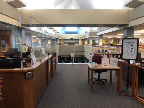 Main Campus Library, right inside entry doors. Shows plexiglass, check in desk, and closed downstairs. Julie Humphrey, Library Director, sits masked at the second staff computer