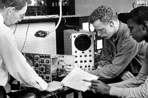 1960s photo of students and instructor looking at insides of outdated electronics equipment with dials