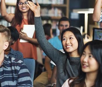 diverse group of students in classroom raising hands to answer a question
