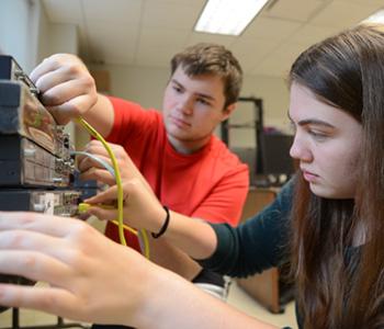 Two students plug cords into ports on the back of a computer