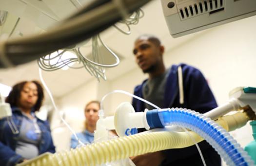 perspective from behind respiratory machine with plastic tubing and three out of focus students in background