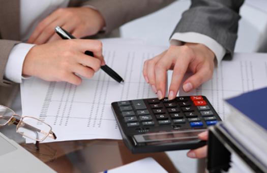 Two business people use a calculator on top of a sheet of numbers.