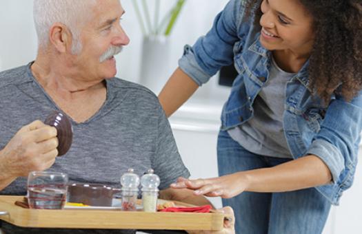 A woman bends towards a man in chair with a meal tray ask if to ask if he needs assistance
