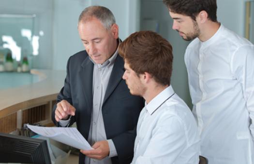 A hotel manager points to a piece of paper as two employees look on.
