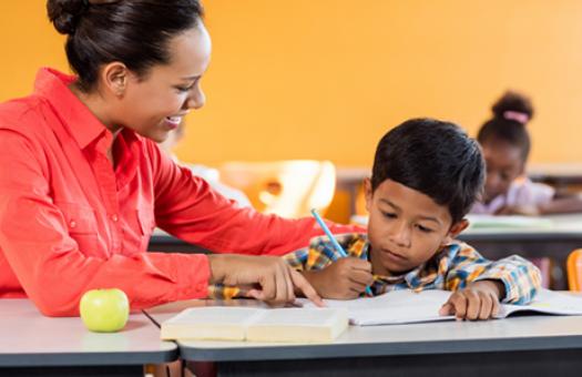 A teacher leans over and points at a book in front of a small child who is staring intently at the page with a pencil in hand.