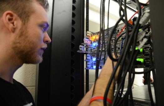 An Information Technology program student looks at a multi-colored server with LED lights as the student uses his hands to work on the server.