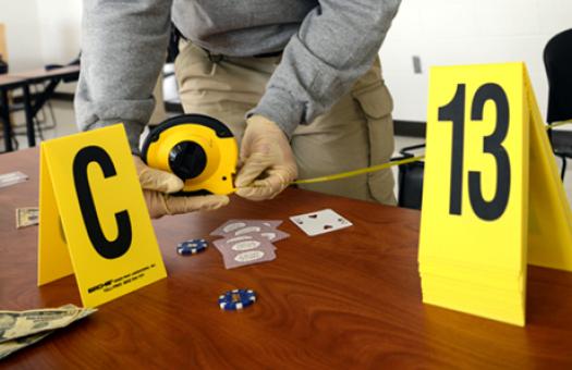 Evidence markers sit on a desk as a Criminal Justice Technology student measures the distance between areas of a training crime scene.