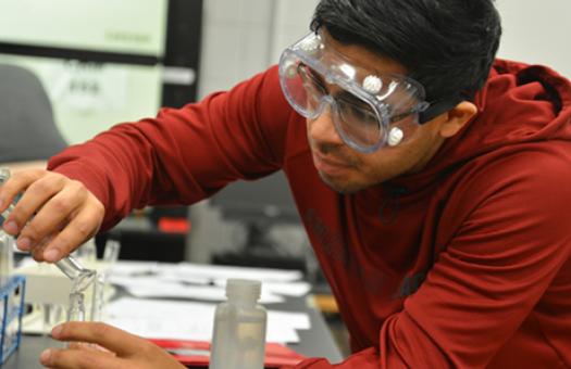 A male student in the Associate in Science program pours liquid into a beaker in a chemistry classroom.