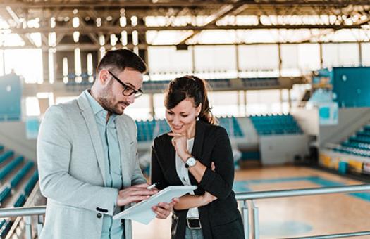 Two individuals wearing professional attire look at a report with gymnasium in background