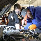 Two students looking under the hood of a car with a flashlight to diagnose a problem