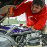 student in red hoodie leaning over car and looking at engine with screwdriver in hand
