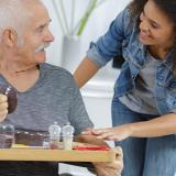 A woman bends towards a man in chair with a meal tray ask if to ask if he needs assistance