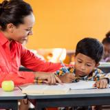 A teacher leans over and points at a book in front of a small child who is staring intently at the page with a pencil in hand.