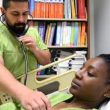 A male student wearing medical scrubs practices using a stethoscope on a female student while she lays on a gurney.