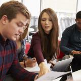 A female student sitting between several other students points to a piece of paper held by one of the male students.