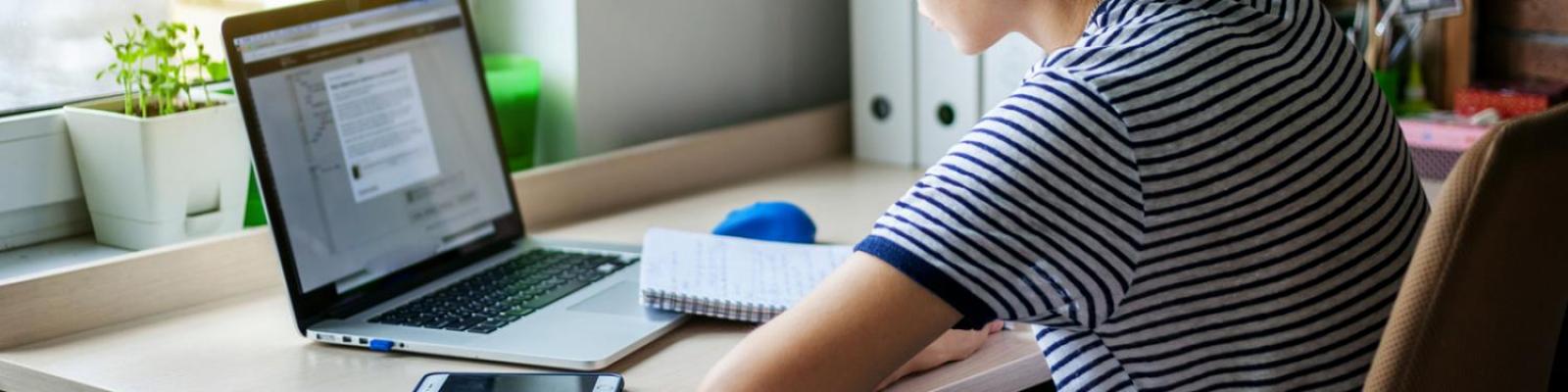 student sitting at desk taking notes while looking at laptop